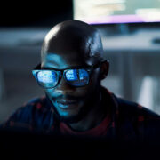 man sitting in dark room facing camera with digital screen in front of him but off screen. wearing black glasses reflecting data on screen in front of him