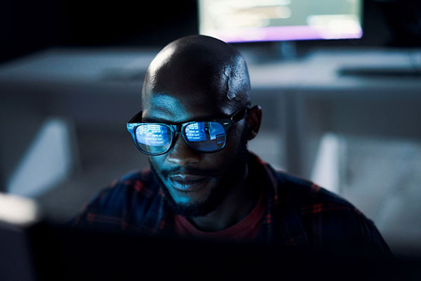 man sitting in dark room facing camera with digital screen in front of him but off screen. wearing black glasses reflecting data on screen in front of him