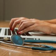 Blue stethoscope lying in front of laptop used by a person to symbolize the presence of technology and cybersecurity in HIPAA and healthcare